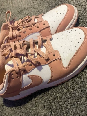  Low rosa och vita sneakers - Säljer ett par Low sneakers i en snygg färgkombination av ljusrosa och vitt. Skorna har klassisk låg siluett, perforerad tåbox och ikoniska swoosh-loggan på sidorna. Ovandelen är i läder och de har matchande rosa skosnören. Perfekta för en trendig streetstyle-look.