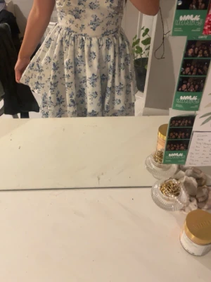 Vit blommig kortklänning med blått - Supersöt vit klänning med blått blommönster och markerad midja. Kjolen är utställd och klänningen är ärmlös, perfekt för varma dagar. Materialet känns lätt och luftigt, vilket gör den bekväm att bära. Aldrig haft på mig den, köptes från plick helt ny fast fanns ingen lapp så jag vet inte vart den är ifrån 💕, skulle vilja gissa boohoo