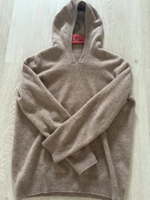 Beige hoodie i merinoull - En stilren beige hoodie i mjuk och skön merinoull. Hoodien har en klassisk huva, ribbade muddar vid ärmslut och nederkant samt en avslappnad passform. Perfekt för dig som gillar en enkel men snygg look.