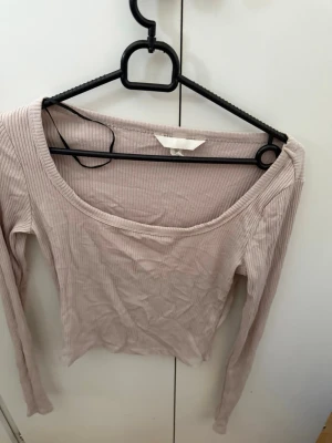 Ljusbeige ribbad långärmad topp H&M - Säljer en ljusbeige långärmad topp från H&M med ribbad struktur och bred, rundad halsringning. Perfekt för att skapa en enkel och snygg look. Materialet är mjukt och stretchigt, vilket gör den bekväm att bära hela dagen.