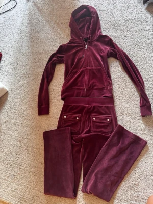 Vinröd velourset från Juicy Couture - Säljer ett vinrött loungewear-set från Juicy Couture i mjuk velour. Setet består av en zip-hoodie med fickor och matchande byxor med fickor och silverfärgade knappar. Perfekt för chill dagar hemma eller när du vill vara extra comfy. 