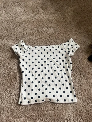Vit prickig offshoulder topp - Snygg vit topp med svarta prickar och offshoulder-design. Kortärmad modell i mjuk bomull, perfekt för en trendig och avslappnad look. Passar dig som gillar lekfulla mönster och vill sticka ut lite extra.