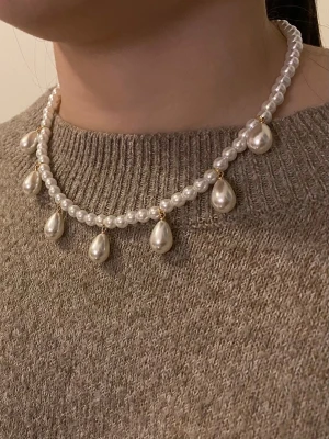 Pärlhalsband med droppformade hängen - Stilrent halsband med små vita pärlor och droppformade pärlhängen. Halsbandet har detaljer i guld mellan pärlorna och ger en klassisk och elegant look. Perfekt accessoar för att lyfta din outfit med en trendig touch.