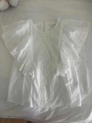 Vit blus med broderade volangärmar - Superfin vit blus med breda volangärmar och broderade spetsdetaljer längs ärmar och framtill. Blusen har rund halsringning och är tillverkad i ett lätt bomullstyg. Perfekt för en fräsch och somrig look.