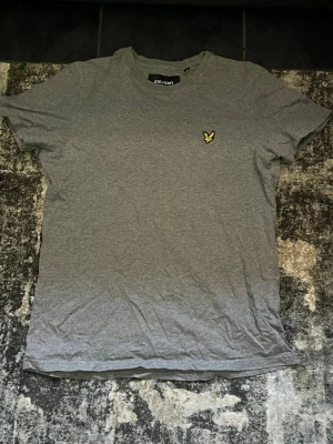 Grå t-shirt från Lyle & Scott - Snygg grå t-shirt från Lyle & Scott med klassisk gul broderad logga på bröstet. T-shirten har rund hals och korta ärmar, perfekt för en clean och enkel stil. Tillverkad i mjuk bomull för skön känsla hela dagen.