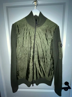 Olivgrön ziptröja från J.Lindberg - Snygg olivgrön cardigan från Jlindberg med stickade ärmar och rygg samt quiltad framsida i blankt material. Hög krage och logopatch på ena ärmen. Perfekt för lager-på-lager och streetwear-stil. Öppen för bytte!