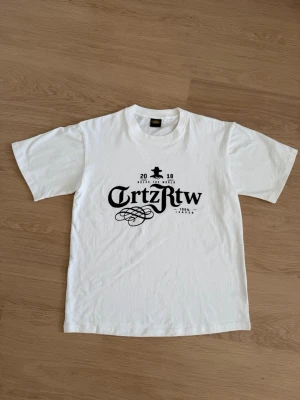 Vit t-shirt med svart tryck Corteiz - Vit t-shirt från Corteiz med svart grafiskt tryck framtill och klassisk rund hals. T-shirten är kortärmad och tillverkad i mjuk bomull, perfekt för en avslappnad och stilren look.