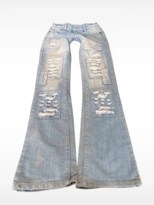Ljusblåa utsvängda ripped jeans - Säljer ett par ljusblåa jeans  med utsvängda ben och rejält slitna detaljer framtill och baktill. Jeansen har klassisk femficksdesign, låg midja och är tillverkade i denim. Perfekta för dig som gillar en edgy och trendig stil. Super snygga, knappen på gylfen saknas som man kan se på bilden men inget som syns och är enkelt att laga!❤️