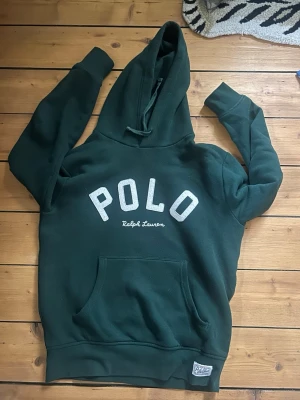 Mörkgrön hoodie från Polo Ralph Lauren - Mörkgrön hoodie med stor vit POLO-text och Ralph Lauren-brodyr på bröstet. Klassisk känguruficka framtill, ribbade muddar och huva. Perfekt för en avslappnad och stilren look. Hör av er vid frågor!