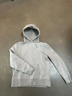 Grå basic hoodie med huva - Enkel och stilren grå hoodie med huva och dragsko. Klassisk känguruficka framtill och ribbade muddar vid ärmslut och nederkant. Perfekt för en avslappnad och casual look. Materialet känns mjukt och bekvämt, troligen bomullsblandning.