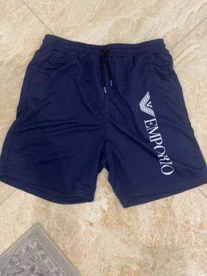 Marinblå shorts från Emporio - Snygga marinblå shorts från Emporio med vit logga längs ena benet. Shortsen har resårmidja med snörning och är gjorda i 100% polyester, vilket gör dem lätta och bekväma. Perfekta för varma dagar eller träning.