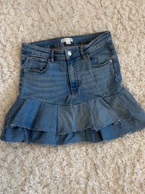 Jeanskjol med volangkant - Egenstudier Snygg blå jeanskjol med volangkant och råa kanter av ett par jeans från gina tricot. Kjolen har klassiska fickor fram och bak samt knapp- och dragkedjestängning. Perfekt för dig som gillar en trendig och avslappnad stil.
