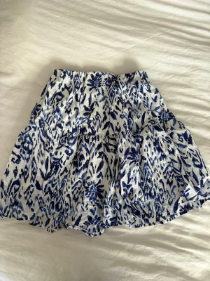 Blåvit kortkjol från Gina Tricot - Kortkjol från Gina Tricot i vitt med blått blommigt mönster. Kjolen har volangdetaljer och resår i midjan för en bekväm passform. Perfekt för varma dagar och enkel att matcha med en enkel topp.