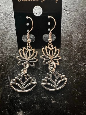 Silvriga lotusörhängen i metall - Säljer ett par snygga silvriga örhängen i metall med dubbla lotusblommor i hängande design. Örhängena har krokar för enkel användning och en stilren, bohemisk känsla. Perfekta för dig som gillar unika smycken med naturinspirerade former.