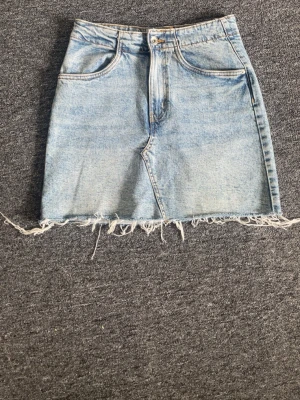 Ljusblå jeanskjol med fransig kant - Trendig ljusblå jeanskjol i kort modell med rå, fransig nederkant. Kjolen har klassiska fickor fram och bak samt dragkedja och knapp framtill. Perfekt för dig som gillar en avslappnad och cool stil.