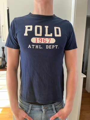 Blå POLO 1967 t-shirt - Mörkblå t-shirt från Polo Ralph Lauren med trycket 'POLO 1967 ATHL. DEPT.' i vitt och rött framtill. Klassisk rund hals och korta ärmar. Perfekt för en avslappnad och sportig stil.