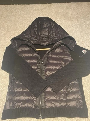 Svart pufferjacka med stickade ärmar Moncler - Snygg svart pufferjacka från Moncler med glansig vadderad front och huva. Ärmarna och sidorna är i stickat material för en cool kontrast. Jackan har dragkedja framtill och Moncler-logga på ärmen. 