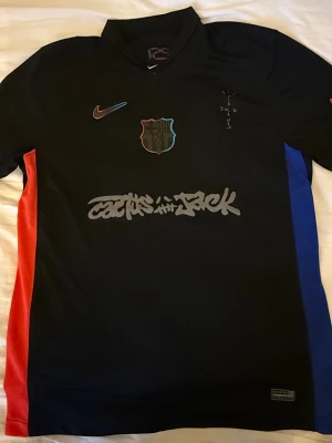 Svart FC Barcelona x Nike matchtröja - Säljer en svart FC Barcelona matchtröja från Nike med klubbmärke och Nike-logga på bröstet. Tröjan har röda och blå paneler på sidorna, grafiskt tryck på framsidan och UNHCR-logga bak. Perfekt för dig som älskar fotboll och streetstyle.