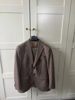 Brun ullkostym från Oscar Jacobson - Snygg brun kostym från Oscar Jacobson i klassisk modell. Kostymen är tillverkad i ull och har två knappar framtill, fickor med lock och en elegant beige insida. Perfekt för dig som vill ha en stilren och tidlös look. Byxor och kavaj!!! Nypris är ca 3-5000