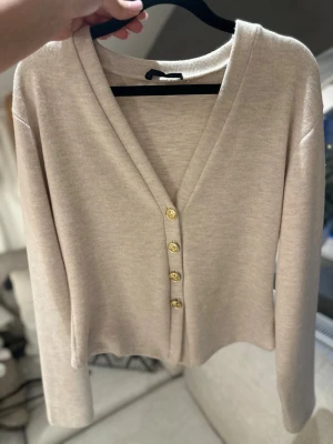Beige kofta från Zara med guldknappar - Stilren beige kofta med djup V-ringning och fyra dekorativa guldknappar framtill. Koftan har lång ärm och är tillverkad i ett mjukt, tunt stickat material. Aldrig använt och är i storlek m från Zara 