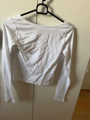 Vit långärmad topp i bomull - En basic vit långärmad topp med en liten djup rygg. Tillverkad i mjuk bomull och har en slim passform som sitter snyggt på kroppen. Perfekt att styla med jeans eller kjol för en clean look.