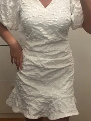 Vit kort klänning med puffärmar - Superfin vit kort klänning med puffärmar och v-ringning. Klänningen har ett diskret blommigt mönster i tyget och volang nertill. Perfekt för student eller sommarens alla tillfällen. Figurnära passform och dragkedja i ryggen.
