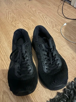 Svarta sneakers från Asics - Säljer ett par helsvarta Asics sneakers med mesh-överdel och syntetdetaljer. Skorna har klassisk snörning och gummisula för bra grepp. Perfekta för dig som gillar stilrena och sportiga skor.