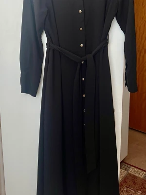 Svart långklänning med bälte - Elegant svart långklänning med långa ärmar, knappar framtill och ett matchande bälte i midjan. Klänningen har en klassisk och stilren design som passar perfekt till många tillfällen.