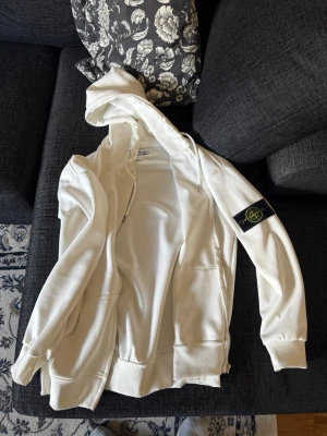 Vit hoodie från Stone Island - Säljer en vit hoodie från Stone Island med dragkedja och klassisk logopatch på vänster ärm. Hoodien har fickor framtill och ribbade muddar vid ärmslut och nederkant. Tillverkad i mjukt bomullsmaterial för en skön och avslappnad look. Storlek m