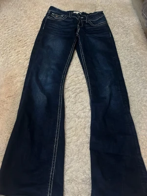 Mörkblå bootcut jeans med kontrastsömmar - Säljer ett par mörkblå bootcut jeans med vita kontrastsömmar och fickor med lock bak från Gina tricot. Jeansen har låg midja och klassisk femficksdesign. Materialet är denim och passformen är avslappnad med utsvängda ben.