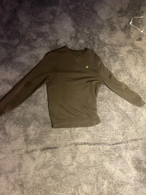 Mörkgrön sweatshirt från Lyle & Scott - Mörkgrön sweatshirt från Lyle & Scott med klassisk rund hals och lång ärm. Tröjan har en liten gul logotyp på bröstet och är tillverkad i mjukt material, perfekt för en avslappnad stil. Igentliten 12-13 är men är lite större skulle jag säga 