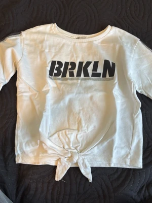 Vit t-shirt med knyt framtill - Vit t-shirt från H&M med svart BRKLN-tryck på bröstet och knytning framtill. T-shirten har korta ärmar och är tillverkad i mjuk bomull, perfekt för en avslappnad streetstyle-look.
