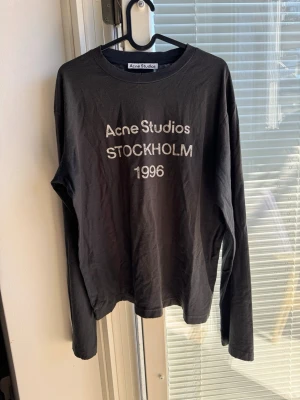 Svart långärmad topp Acne Studios - Svart långärmad topp från Acne Studios med vit text 'Acne Studios STOCKHOLM 1996' på bröstet. Klassisk rund halsringning och avslappnad passform. Perfekt för en stilren och trendig look.