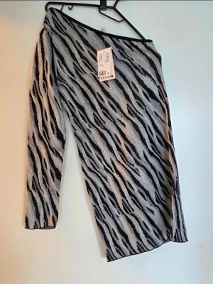 Zebramönstrad oneshoulder klänning med prislapp - Snygg zebramönstrad klänning med oneshoulder-design och lång ärm. Klänningen har svart och grått mönster och är tillverkad i ett stretchigt material som sitter tight. Perfekt för dig som vill sticka ut med en trendig look.