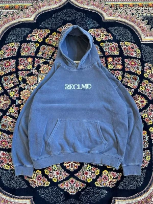 Blå hoodie från RECLMD med tryck - Säljer en blå hoodie från RECLMD med vit logga framtill och stort vintageinspirerat tryck på ryggen. Hoodien har en klassisk känguruficka och huva. Perfekt för dig som gillar streetwear och unika detaljer.