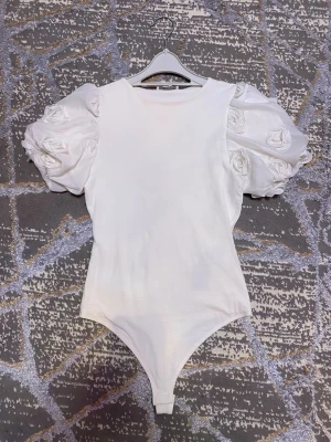 Vit body med puffärmar från Amisu - Trendig vit body från Amisu med unika puffärmar dekorerade med tygrosor. Kroppsnära passform och rund halsringning. Perfekt för dig som vill sticka ut med en cool och romantisk look. Tillverkad i mjukt material för extra komfort.
