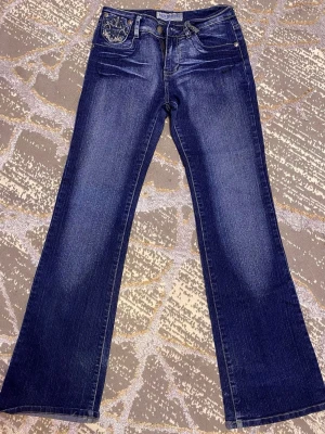 Blå bootcut jeans med broderade fickor  - Snygga blå bootcut jeans från Run Sheng med slitningar och broderad detalj på fickan. Jeansen har klassisk femficksdesign och är tillverkade i denim med lätt stretch. Perfekta för dig som gillar en retroinspirerad look med lite extra attityd. Den är lite sliten längst ner på en ben bara, men den syns inte. Köpte för 600kr. 