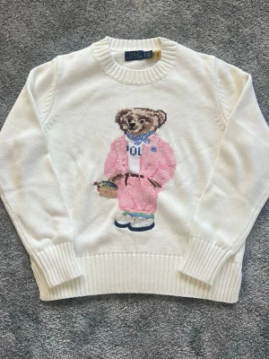 Klassisk stickad Polo Ralph Lauren Bear tröja – Vit/Benvit - Vit stickad tröja från Polo Ralph Lauren med ikonisk björn i rosa outfit på framsidan. Tröjan har rund halsringning och ribbade muddar vid ärmslut och nederkant. Perfekt för dig som gillar unika och lekfulla detaljer i din stil. Längd 56cm och använda fåtal gånger!
