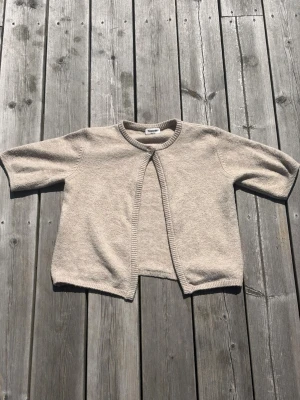 Beige kortärmad kofta  - Stilren beige kofta från Kappahl med korta ärmar och öppen front. Koftan är stickad i mjukt material och har ribbade kanter vid hals, ärmslut och nederkant. Perfekt för lager-på-lager och enkel att matcha med olika outfits.