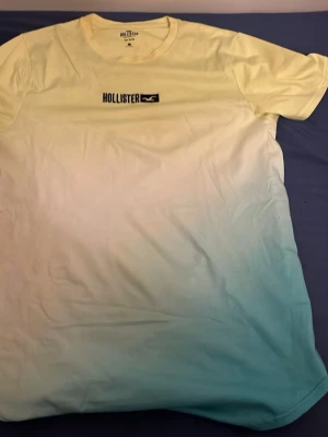 Gul och blå ombre t-shirt Hollister - Snygg t-shirt från Hollister i 100% bomull med ombre-färgskiftning från ljusgul upptill till ljusblå nedtill. Klassisk rund halsringning och tryckt Hollister-logga på bröstet. Perfekt för en avslappnad och trendig stil.