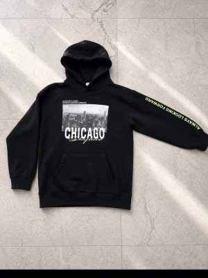 Svart hoodie med Chicago-tryck H&M - Svart hoodie från H&M med stor vit text 'CHICAGO' och stadsmotiv på bröstet. På ena ärmen står det 'ALWAYS LOOKING FORWARD' i grönt. Klassisk känguruficka och huva. Perfekt för en cool streetstyle-look.
