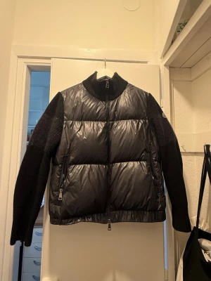 Svart pufferjacka från Moncler - Snygg svart pufferjacka från Moncler med glansig vadderad front och stickade ärmar. Jackan har hög krage, två dragkedjefickor och Moncler-logga på ärmen. Perfekt för kalla dagar och ger en cool streetstil. Ett litet hål längst ut vid armen. Det står att den är i storlek 40 men den passar mig perfekt och jag är 36/38 vanligtvis
