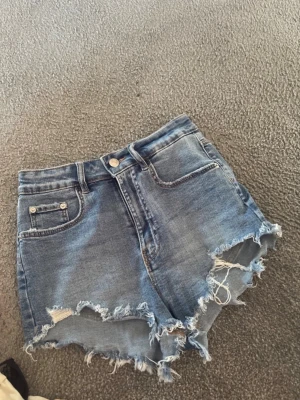 Blå jeansshorts med fransar från Zara - Säljer ett par blå denimshorts från Zara med hög midja och råa, fransiga kanter. Shortsen har klassisk femficksdesign och slitna detaljer för en cool och avslappnad look. Perfekta för sommardagar och festivaler.