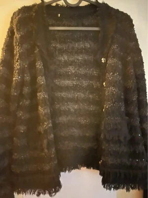 Svart glittrig kofta med struktur - Säljer en svart kofta med glittriga detaljer och tydlig struktur. Koftan har lång ärm och är tillverkad i ett fluffigt, texturerat material med inslag av paljetter som ger extra glans. Perfekt för dig som gillar att sticka ut med din stil.