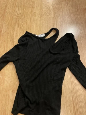 Svart ribbad oneshoulder topp NA-KD - Trendig svart ribbad topp från NA-KD i oneshoulder-modell. Toppen har lång ärm på ena sidan och är av mjuk, stretchig kvalitet. Perfekt för dig som vill sticka ut med en cool och modern look.