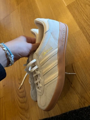 Adidas Gazelle vita sneakers - Snygga Adidas Gazelle sneakers i vitt/ljusblå mocka med klassiska tre ränder på sidan och gummisula i ljusbrunt. Guldfärgad Gazelle-text på sidan och Adidas-logga på hälen. Skorna har lågt skaft och vita skosnören. Perfekta för en clean och stilren look.