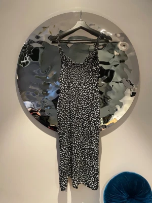 Svart blommig långklänning - Svart långklänning med vita blommor och smala axelband. Klänningen har en lös passform och är tillverkad i ett lätt material som ger ett snyggt fall. Perfekt för dig som gillar en avslappnad men trendig stil.