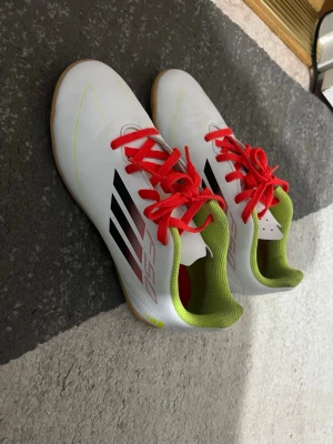 Adidas vita fotbollsskor med röda snören - Säljer ett par snygga vita Adidas fotbollsskor med röda snören och svarta detaljer på sidan. Insidan är limegrön och yttermaterialet är syntetiskt för bra bollkontroll. Perfekta för inomhusspel och har en klassisk Adidas-design.