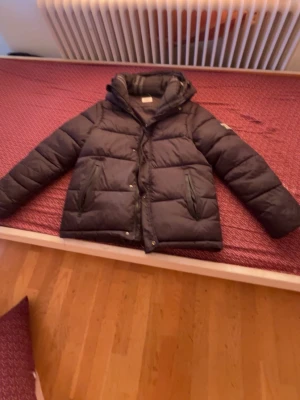 Svart dunjacka från Burberry - Snygg svart dunjacka från Burberry med huva och tydlig logga på ärmen. Jackan har två framfickor med dragkedja och klassisk quiltad design. Perfekt för kalla vinterdagar och riktigt varm tack vare dunfyllningen.den kan även göras till en väst.