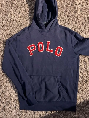 Marinblå hoodie från Polo Ralph Lauren🤩🙌 - Snygg marinblå hoodie från Polo Ralph Lauren med stor röd POLO-text i frotté på bröstet. Klassisk känguruficka framtill och huva. Perfekt för en avslappnad streetwear-look. Tillverkad i mjuk bomullsblandning.🤩🙌